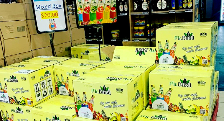 Stacks of yellow boxed beverage cartons labeled 'Mr. Basil' in a store aisle, with a sign reading 'Mixed Box $20.00' displayed above.