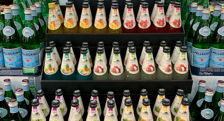 Bottles of flavored drinks with fruit labels are neatly arranged on supermarket shelves; San Pellegrino bottles are visible in the background.