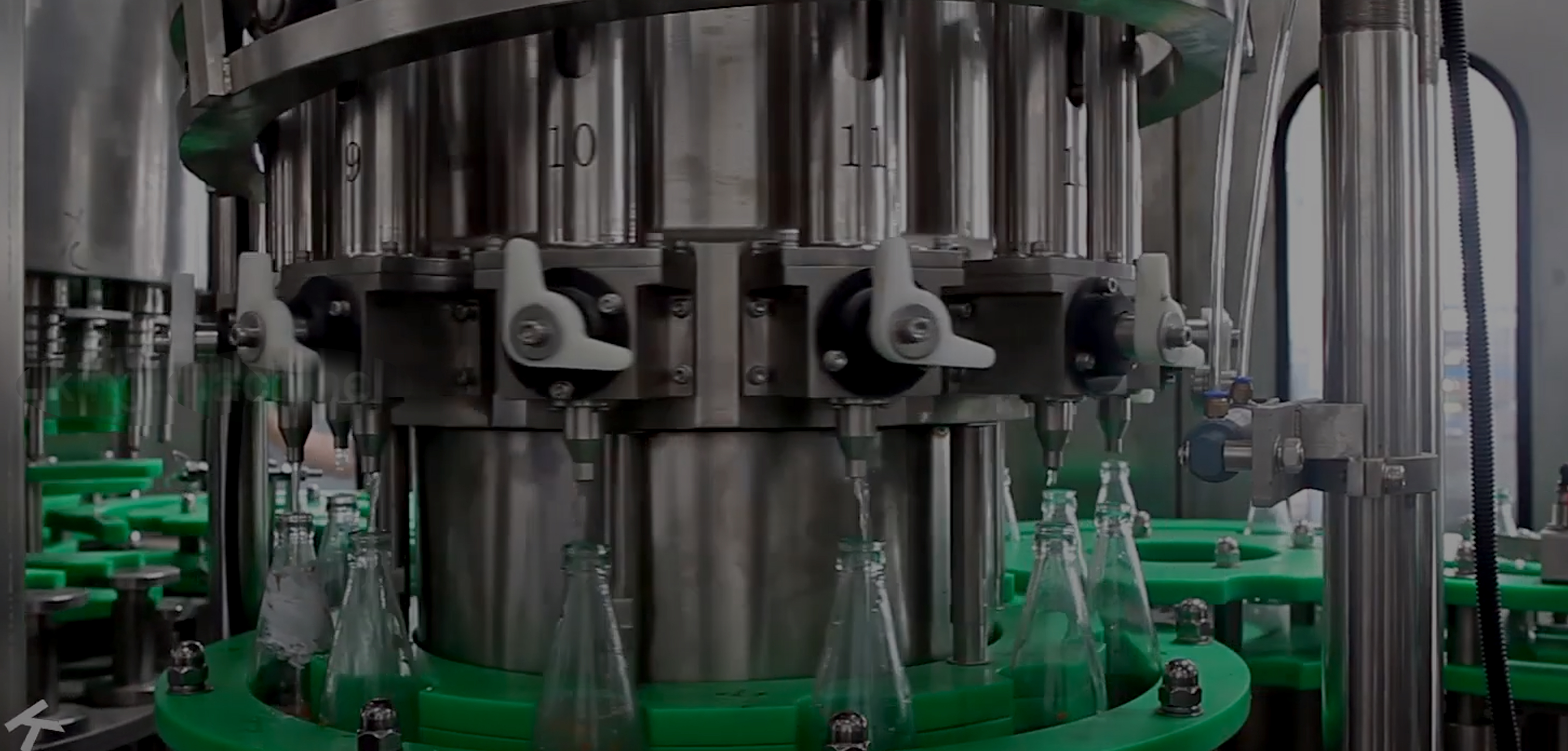 An industrial bottling machine filling clear bottles with green liquid, set against a dark background with moving conveyor belts.